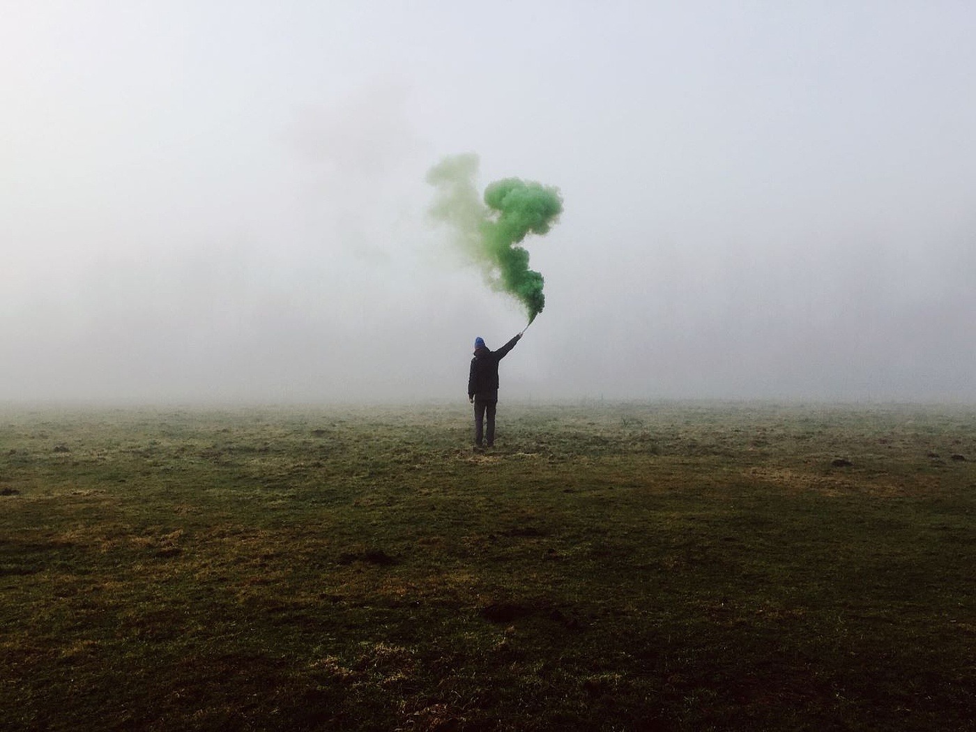 Junger Mann steht mit einer grünen Rauchfackel mit dem Rücken zur Kamera auf einem vernebelten Feld